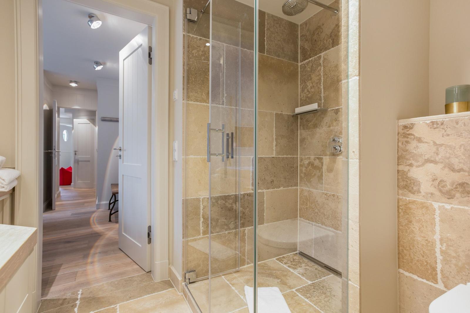 Shower with glass enclosure and stone tiles. View into hallway with wooden floor.