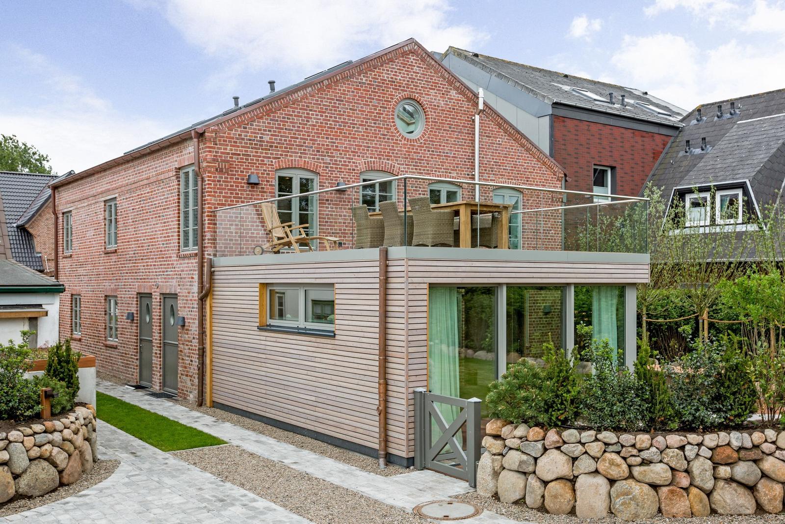 Red brick house with balcony and wooden cladding. Garden with stone wall and plants.