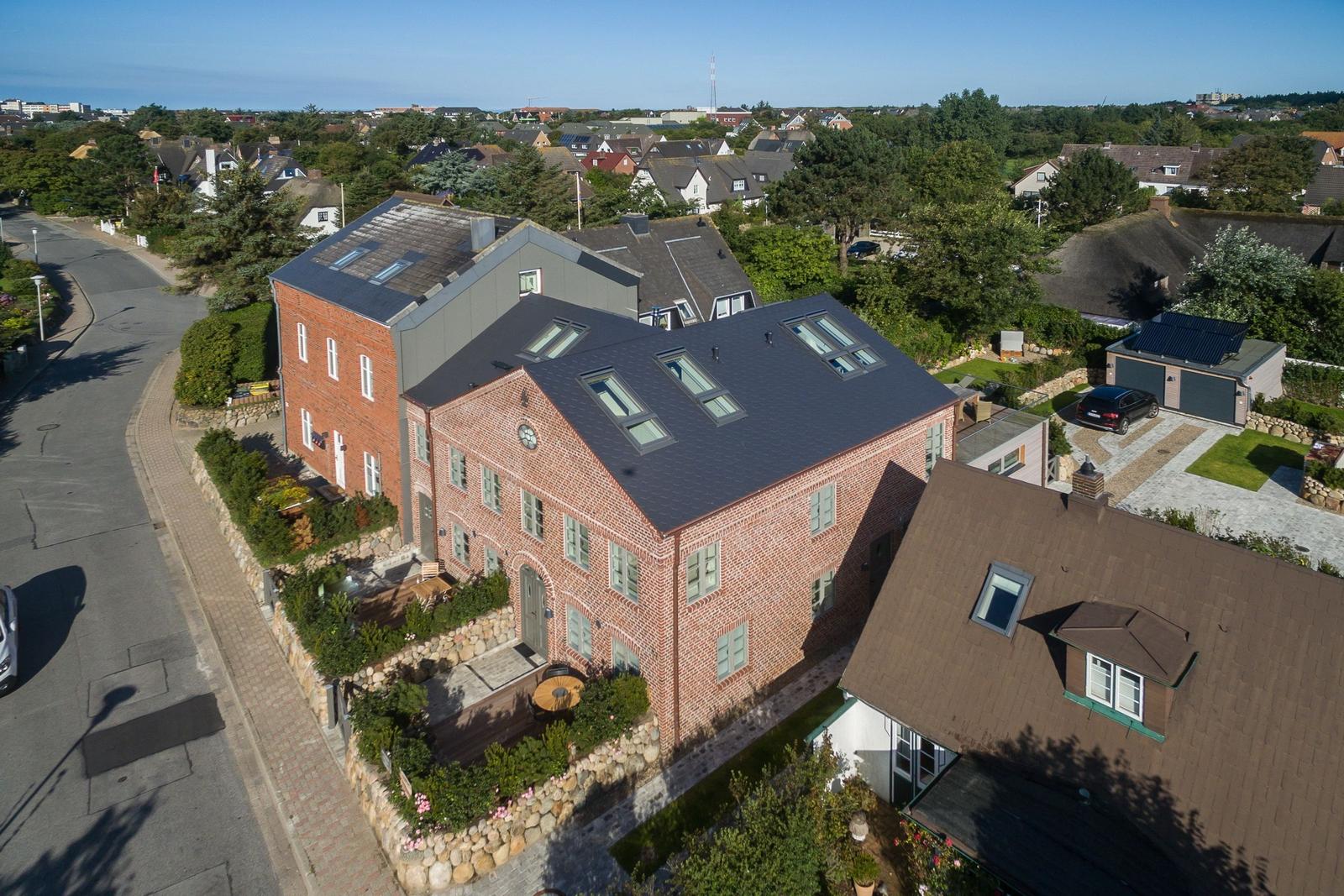 Red brick house with dark roof and multiple skylights. Surrounded by greenery and a paved path.