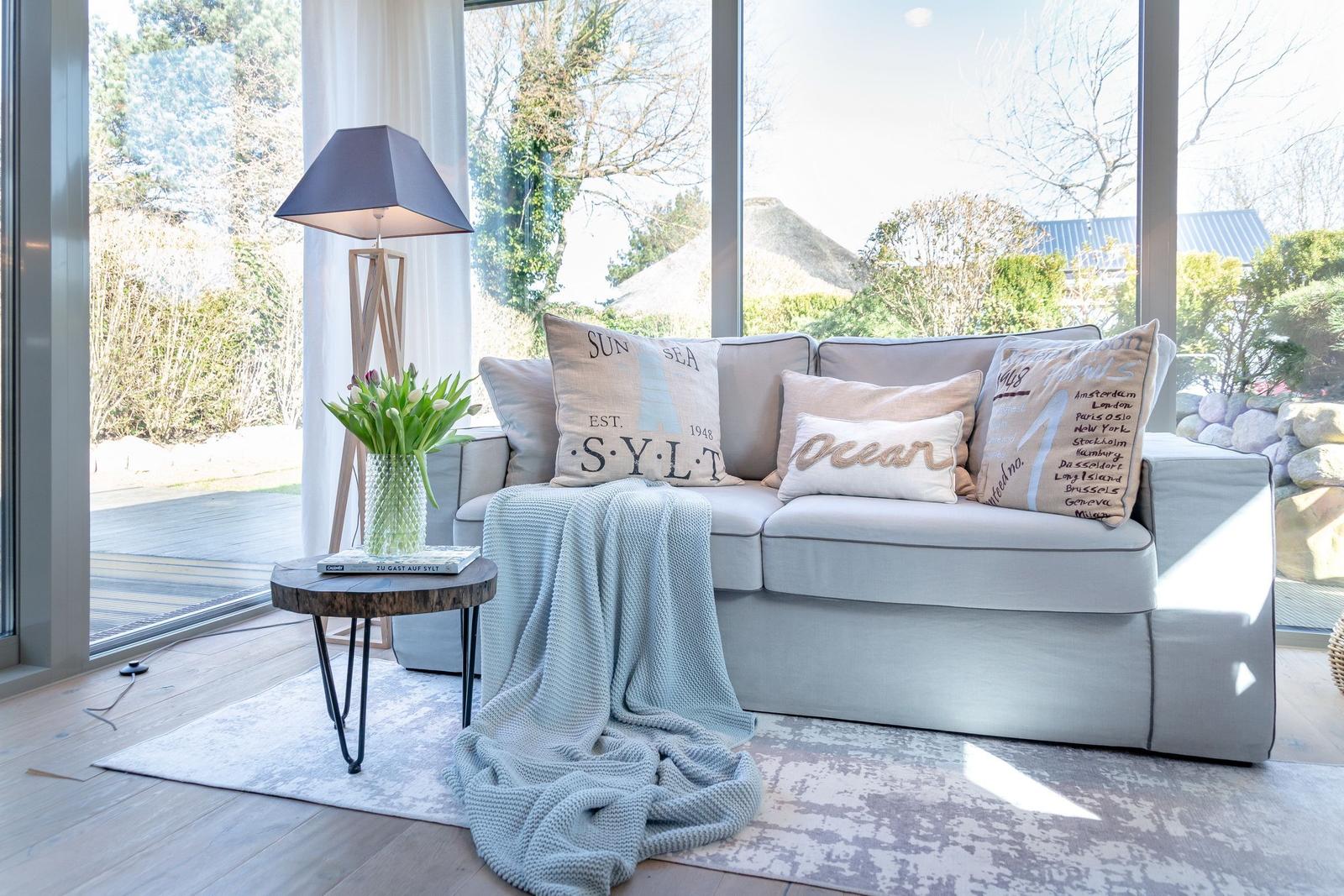 A sofa with cushions and blanket in front of large windows. Table with flowers and lamp beside it.