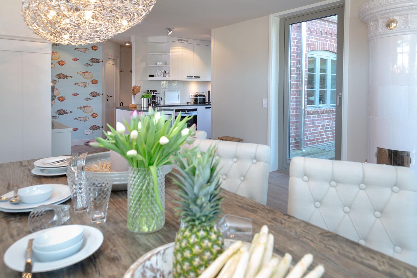 Dining table with chairs, flowers, and fruit. Kitchen and hallway in background.