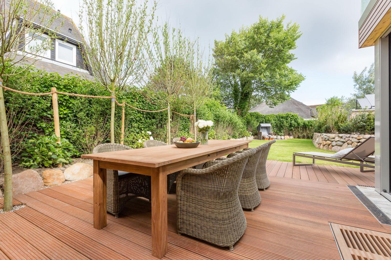 Wooden table with chairs on terrace. Green areas and trees in background.