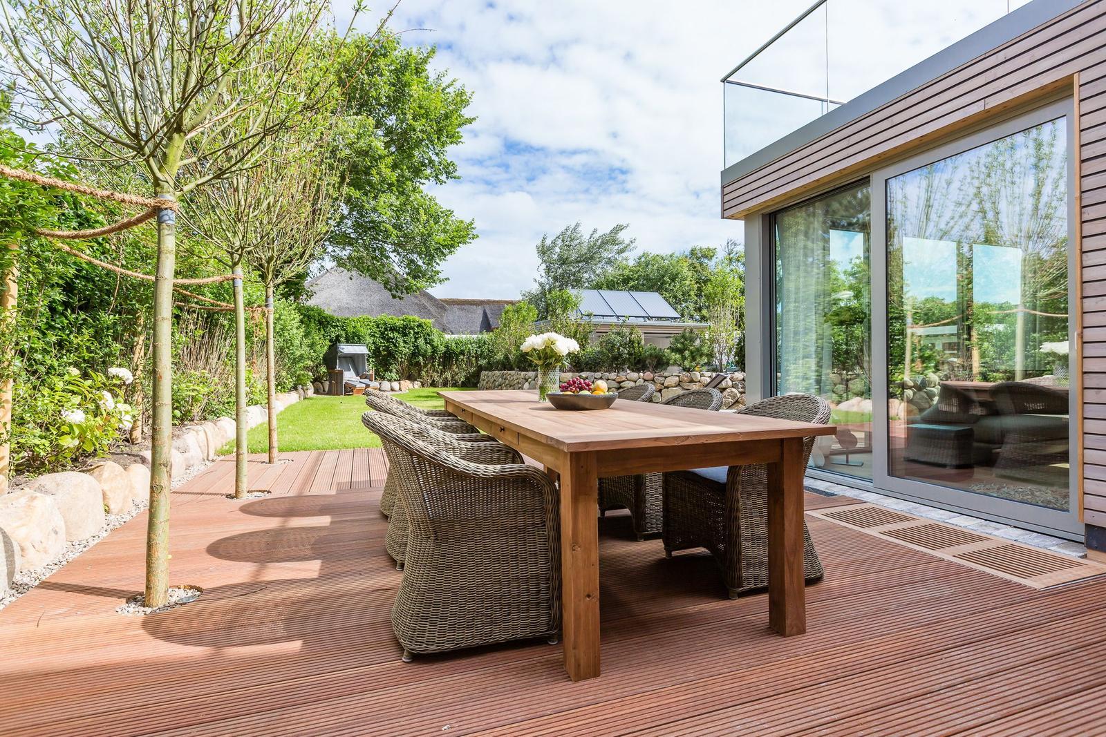 Wooden table with chairs on terrace beside modern house with glass facade.