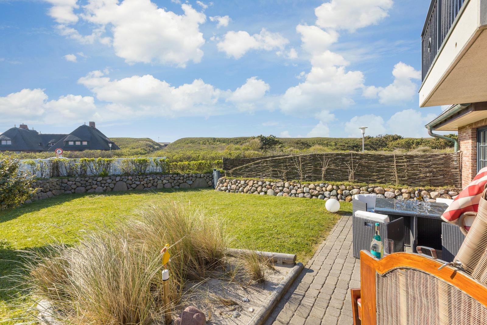 Terrasse mit Sitzgelegenheiten und Blick auf grünen Garten und Strandhaus im Hintergrund.