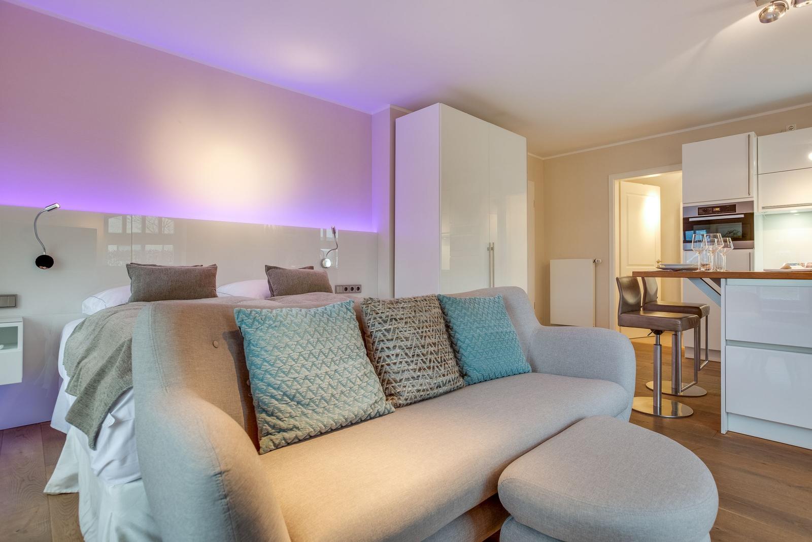 Bedroom with sofa and modern kitchen area in the background.