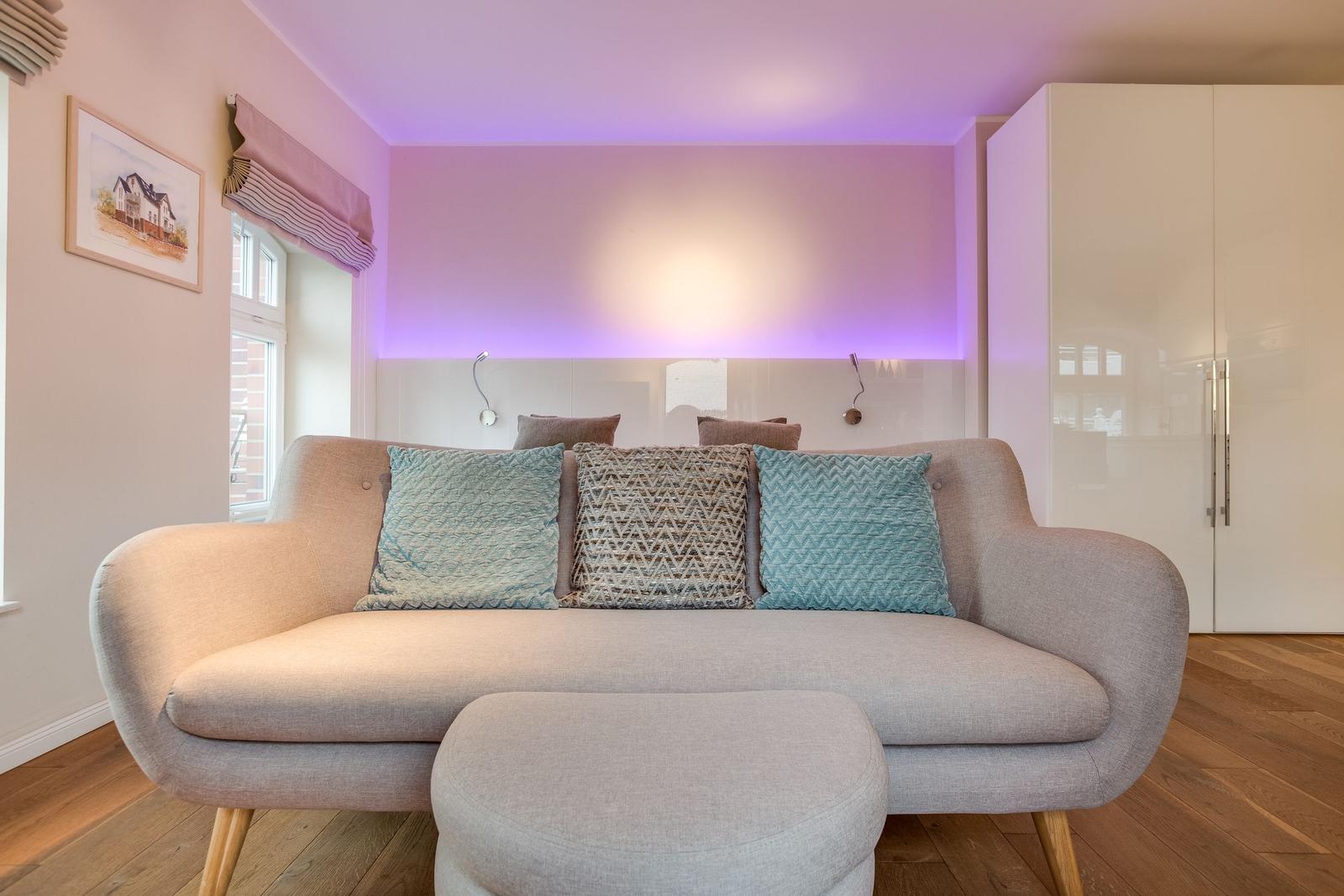 A sofa with cushions in front of a wall with LED lighting and a window.