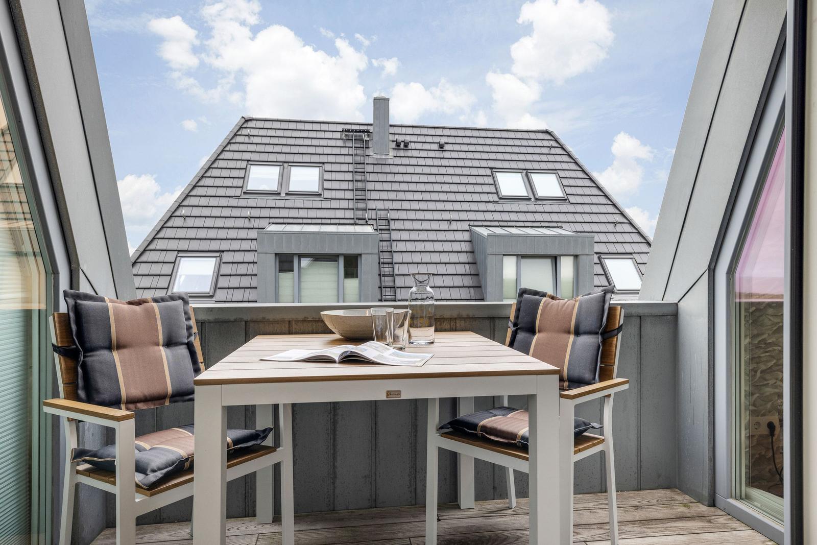 Terrasse mit Tisch und zwei Stühlen. Blick auf ein Haus mit Schiebedachfenstern.