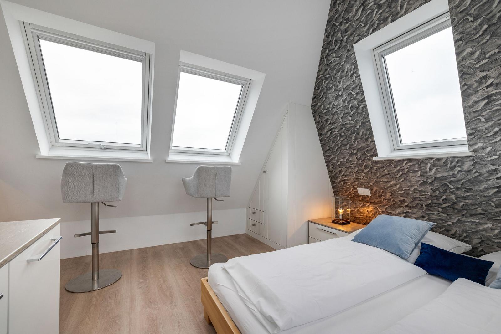 Schlafzimmer mit Dachschrägen, drei Fenstern und einem Bett mit Kissen.