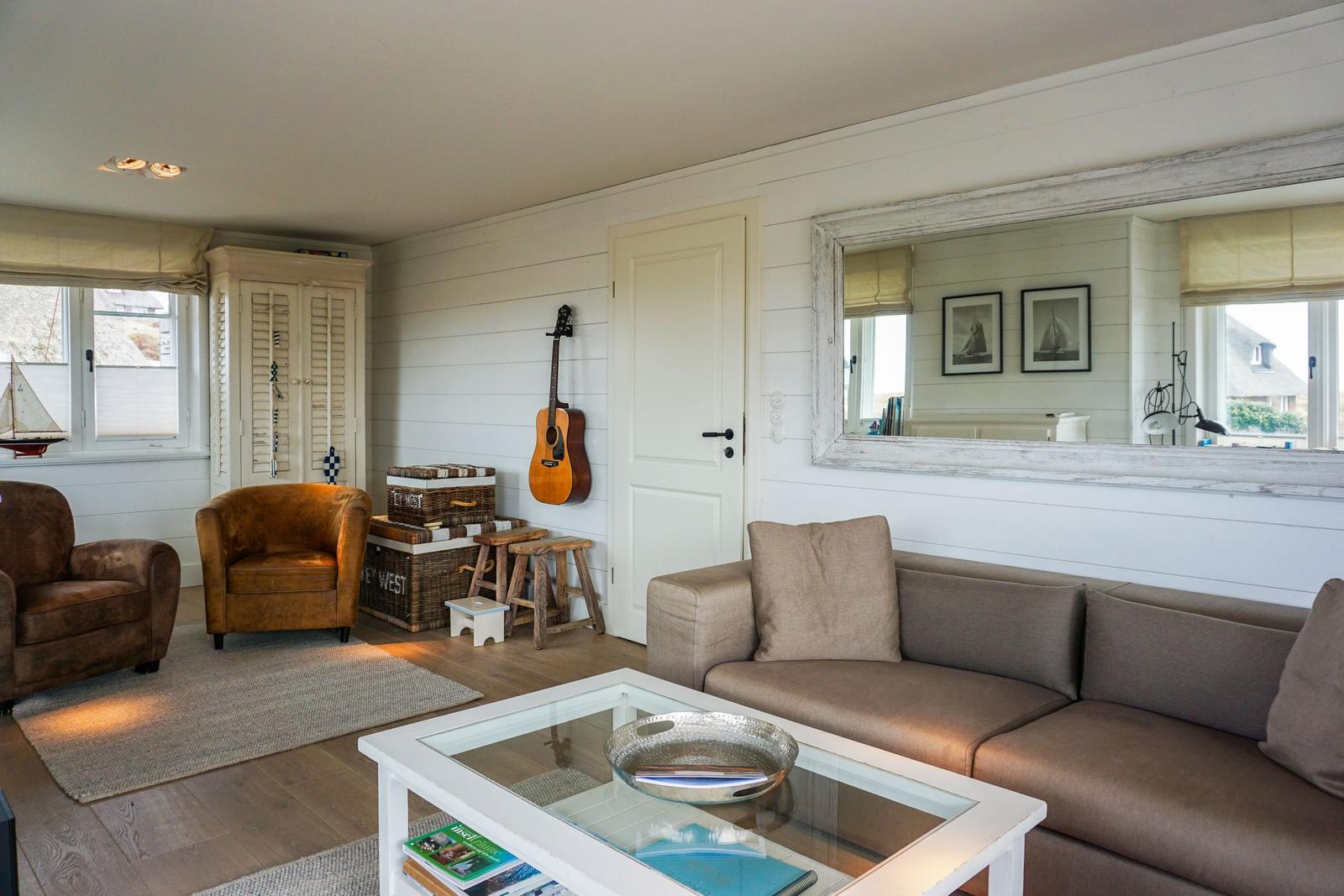 Wohnzimmer mit Sofa, Sessel, Glas-Tisch und Akustikgitarre an der Wand.