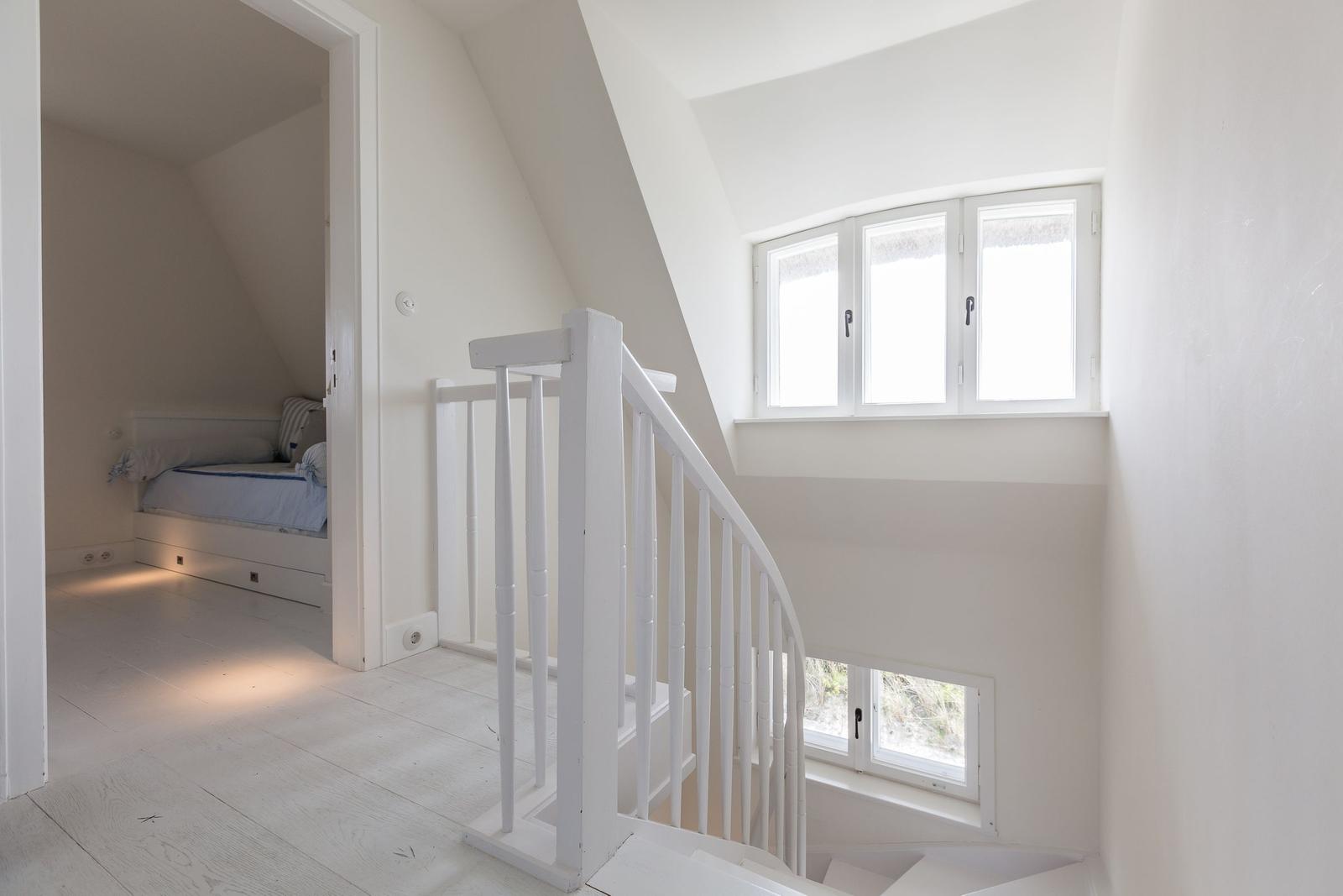 Treppenhaus mit weißer Treppe und Fenster. Im Hintergrund ist ein Schlafzimmer mit Bett sichtbar.