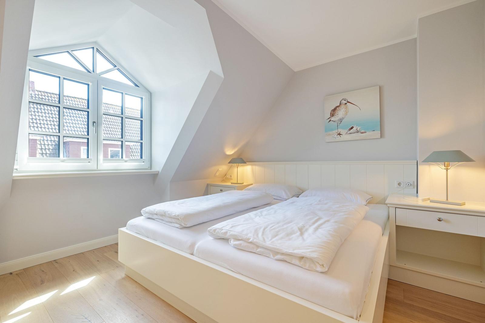 Schlafzimmer mit Dachschrägen, großem Fenster und Bett mit weißen Laken.