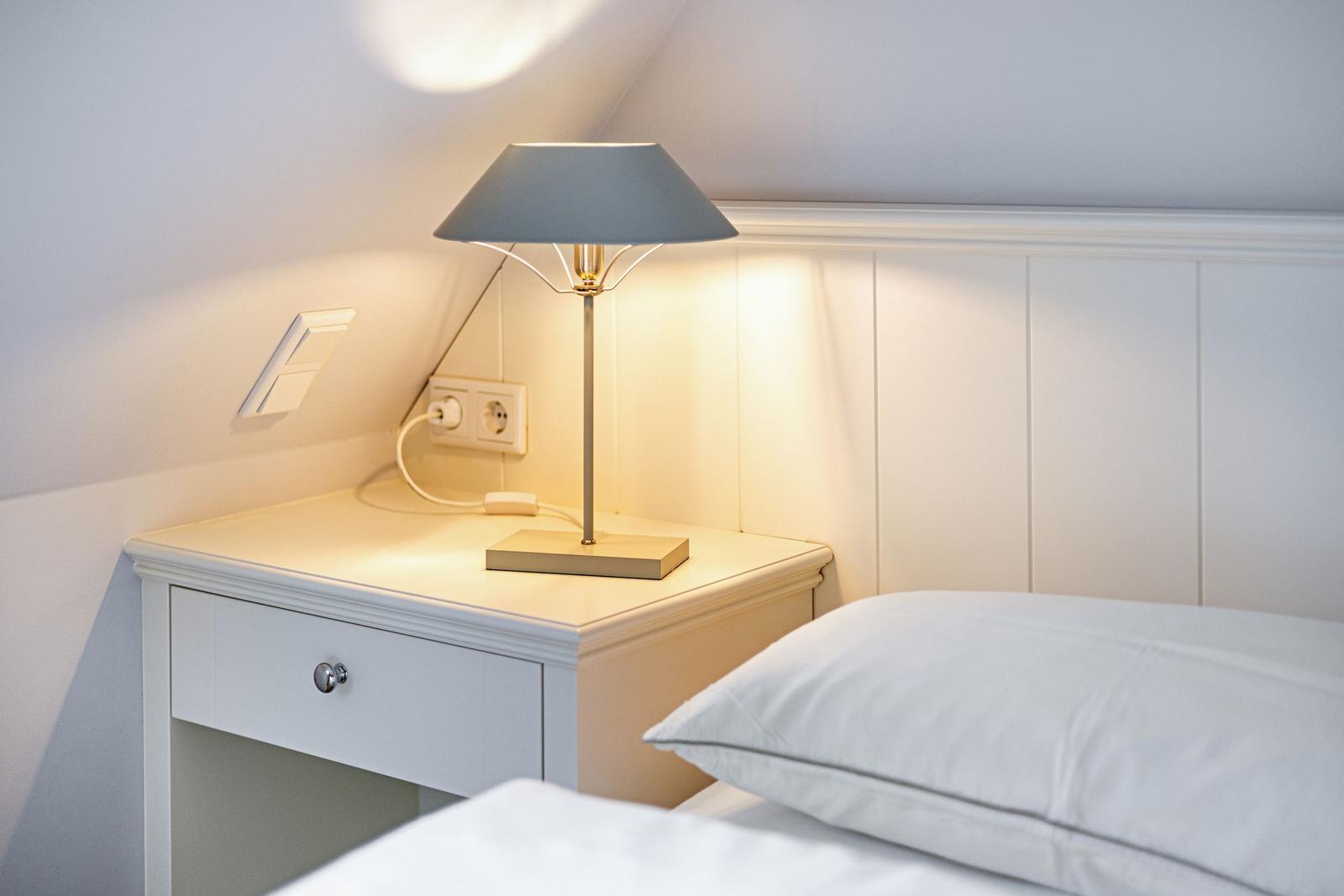 Schlafzimmer mit Nachttisch, Lampe und Bett. Wandverkleidung und Steckdose sichtbar.