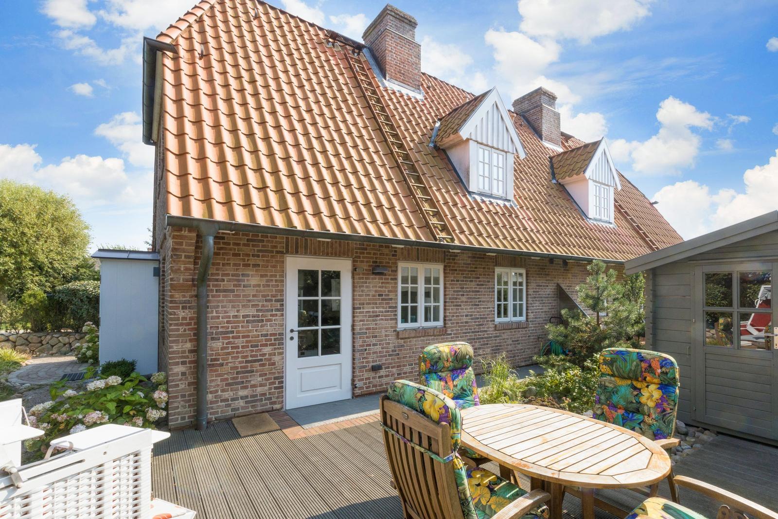 Haus mit Ziegeldach, Terrasse und Gartenmöbel unter blauem Himmel.