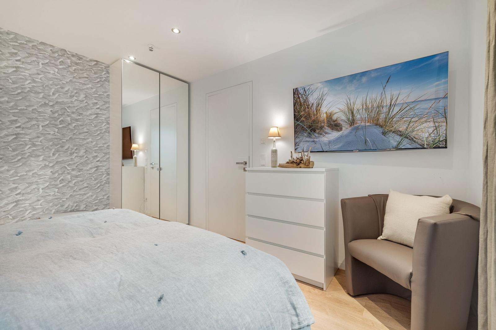 Schlafzimmer mit Bett, Schrank, Kommode und Sessel. Wandbild zeigt Strand.