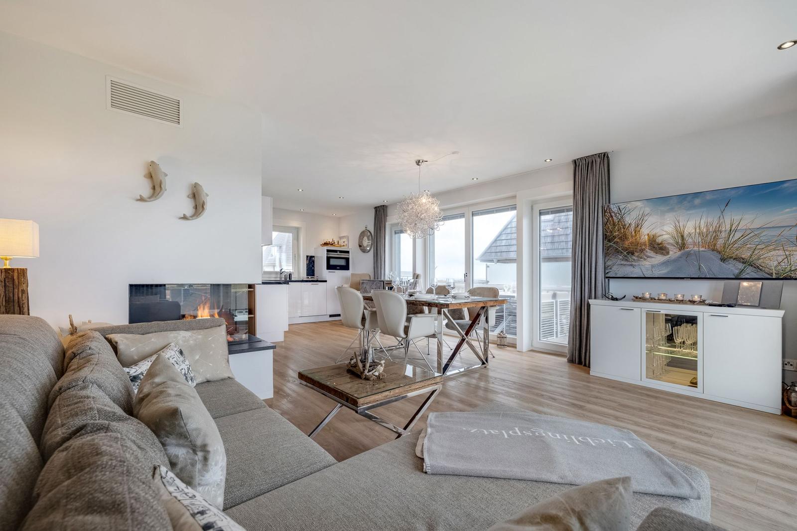 Wohnbereich mit Kamin, Esstisch und TV-Schrank. Blick auf Terrasse und Strand.