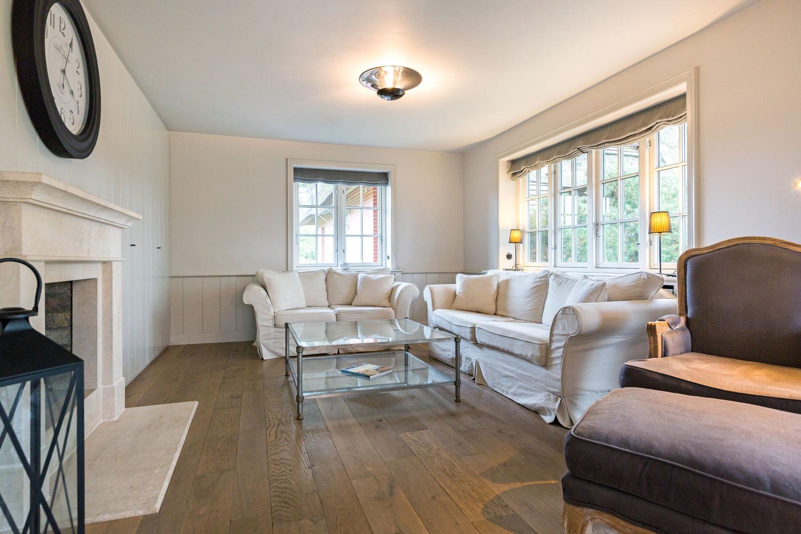 Wohnzimmer mit zwei Sofas, Glas-Tisch und Kamin. Holzboden und große Fenster.
