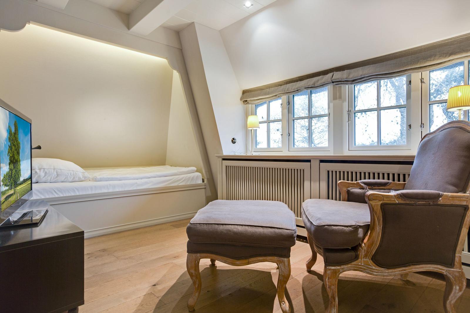 Zimmer mit Bett, Sessel und Fenster. Holzboden und Fernseher.