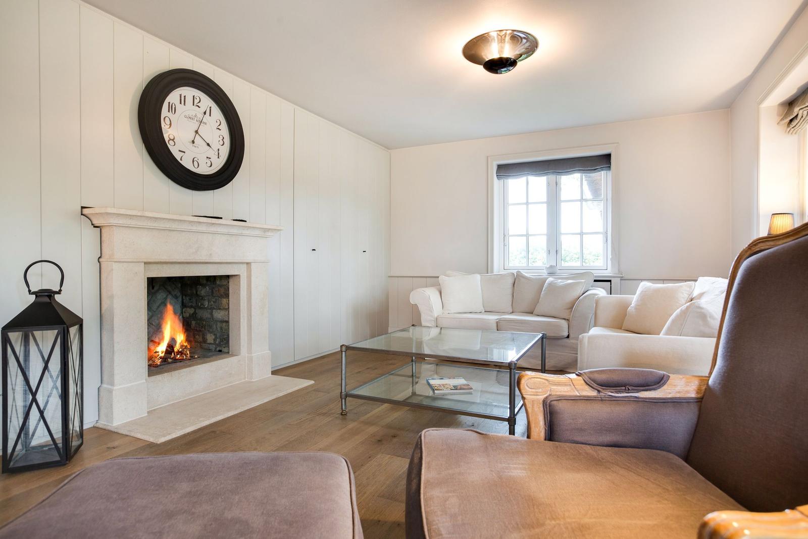 Wohnzimmer mit Kamin, Sofa und Glas-Tisch. Wanduhr und Fenster sichtbar.