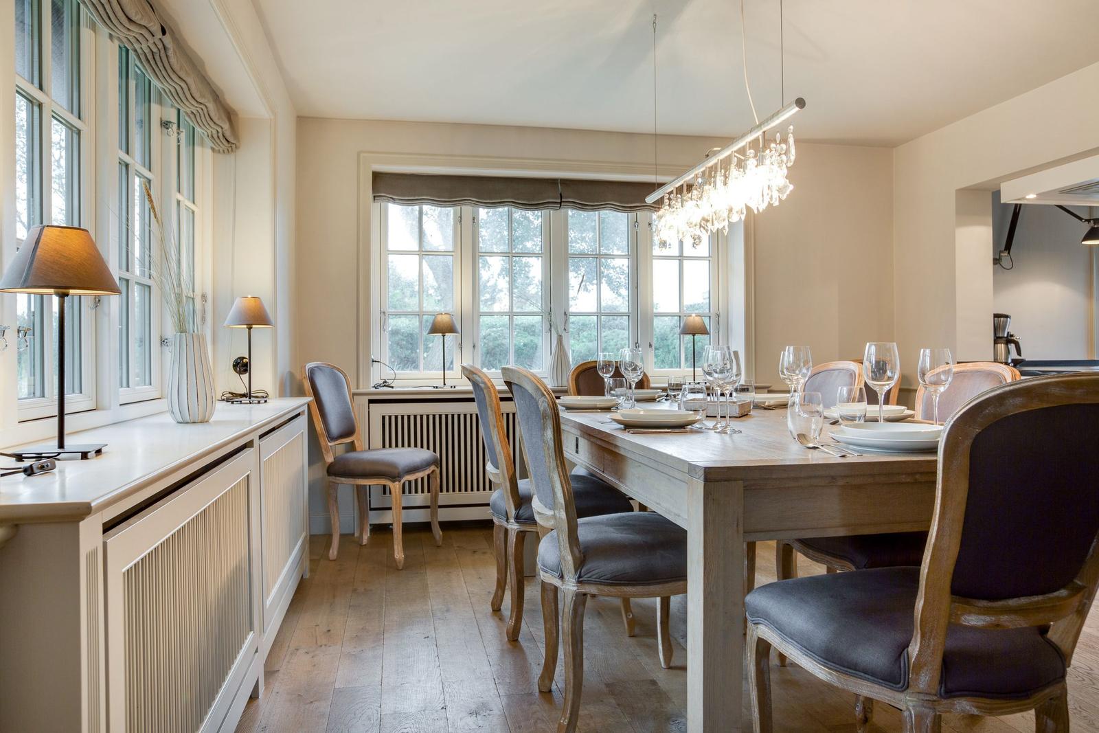 Esszimmer mit Holztisch, Stühlen und Fenstern. Deckenleuchte und Bodenfliesen.