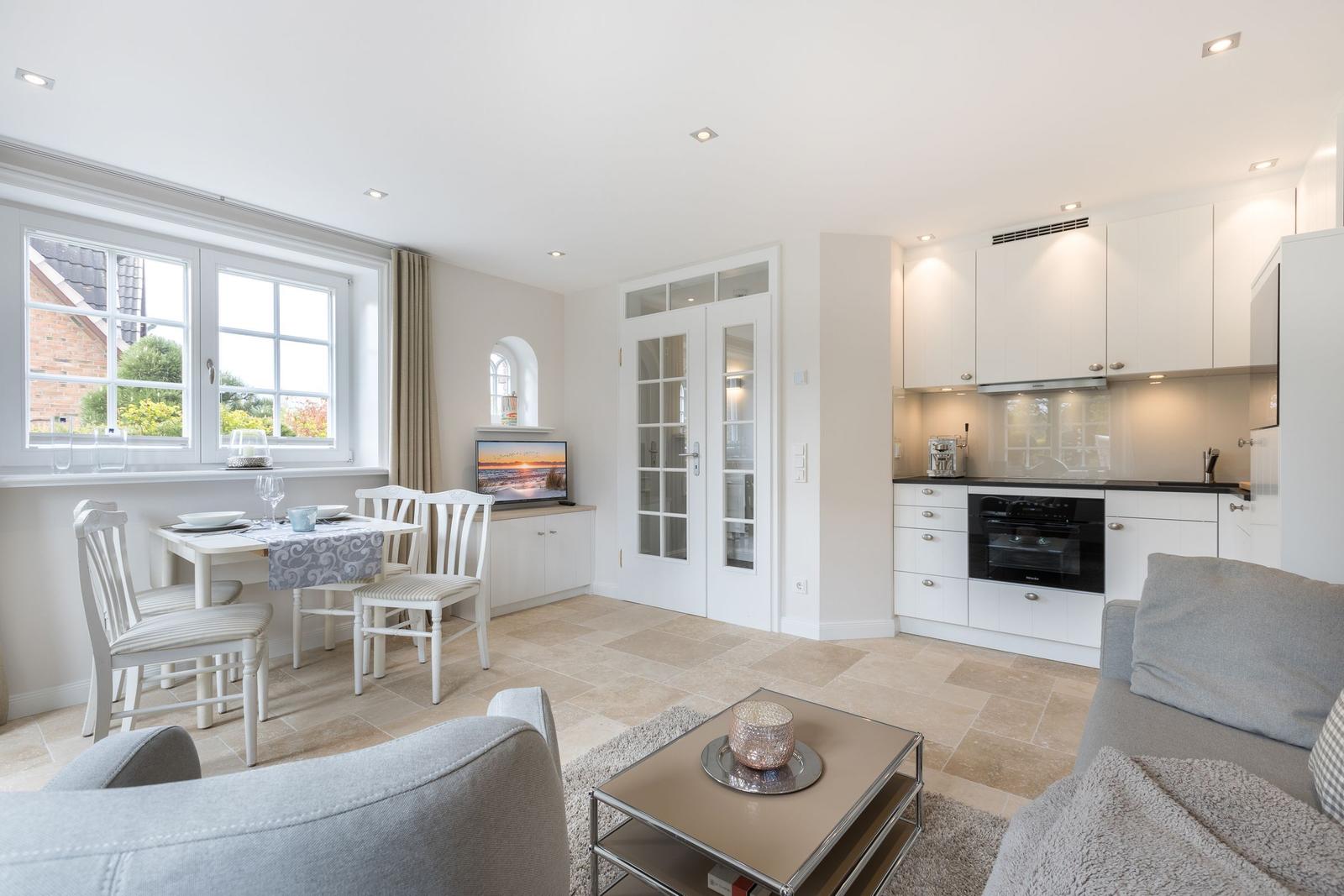 Living kitchen with dining area, sofa, and TV. White furniture, large windows.