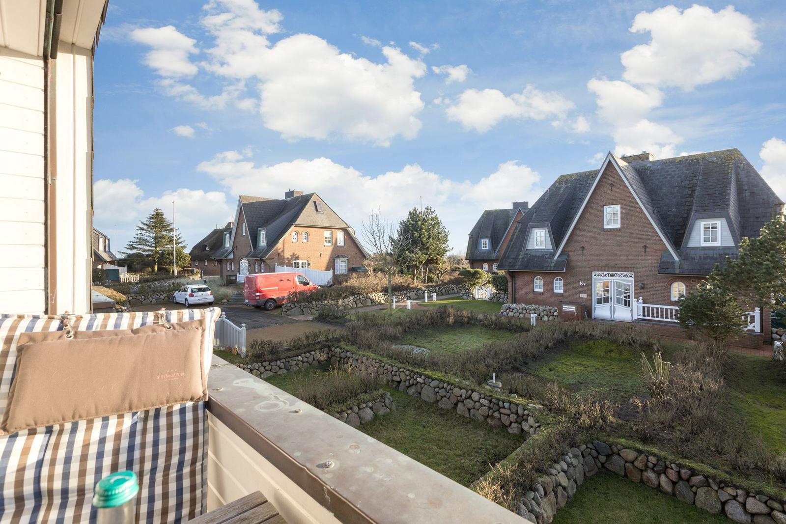 Balcony view of brick houses with gardens and stone walls under blue sky.