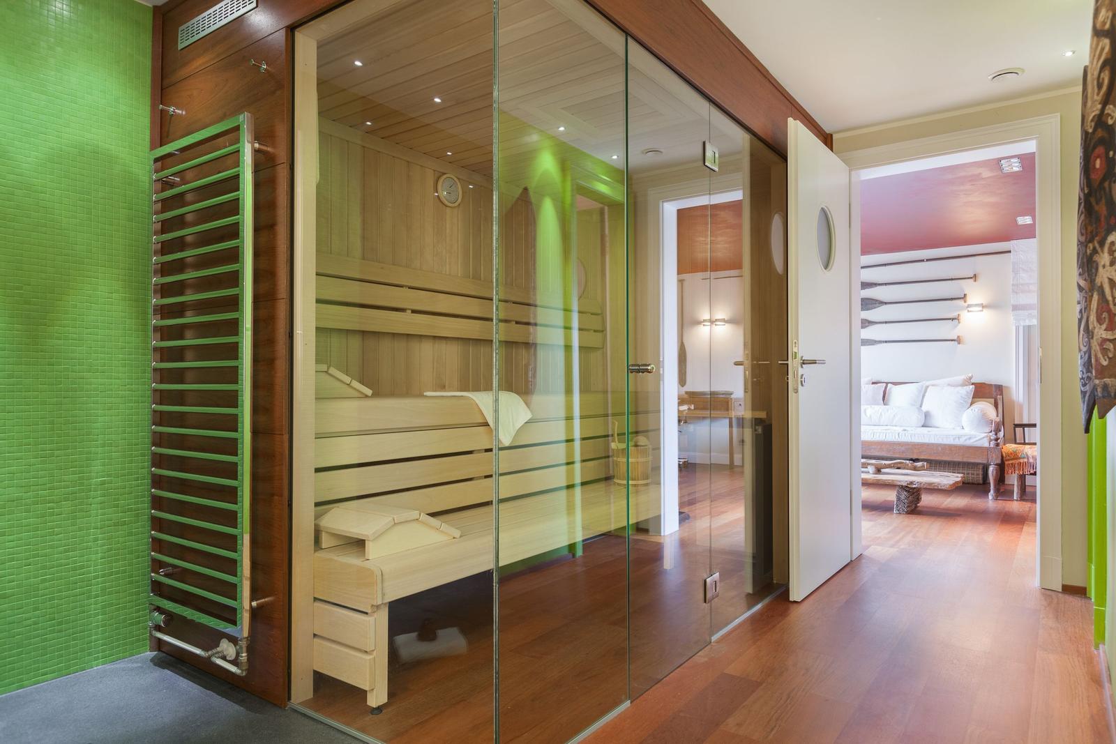 Sauna mit Glaswand, Holzbänken und grüner Wand. Blick in Schlafzimmer mit Bett.