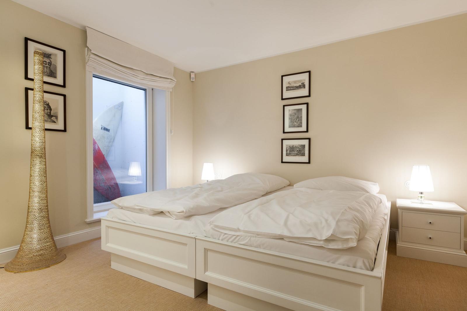 Schlafzimmer mit weißem Bett, Nachttisch und Fenster mit Surfbrett.