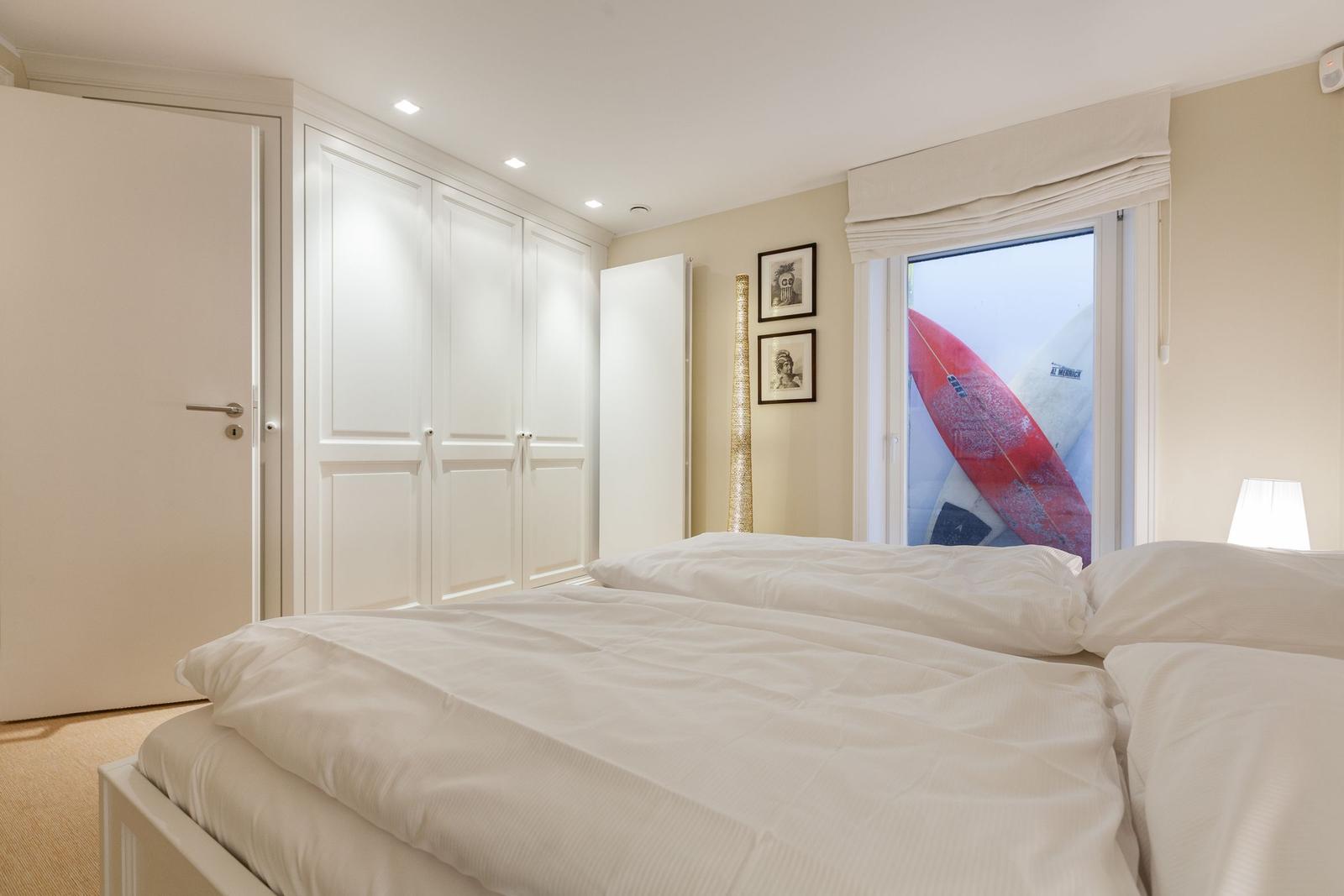 Schlafzimmer mit weißem Bett, Schwebetüren und Fenster mit Surfbrettern.