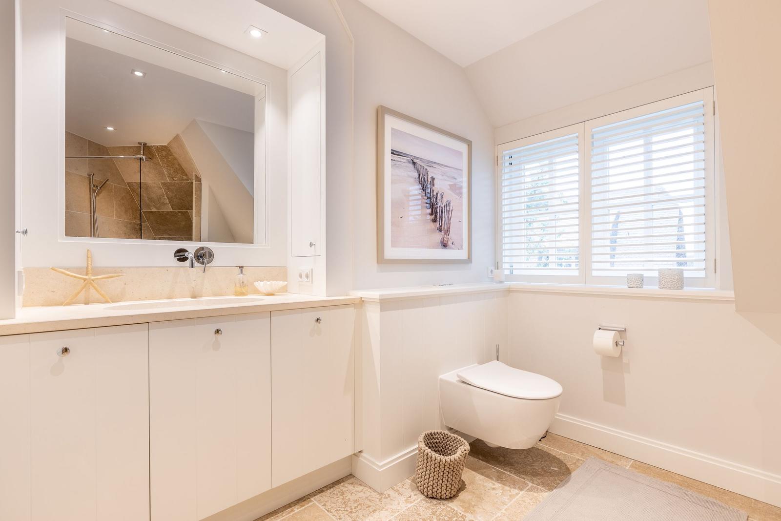 Badezimmer mit Waschbecken, Toilette und Spiegel. Fenster mit Jalousien und Wandbild.