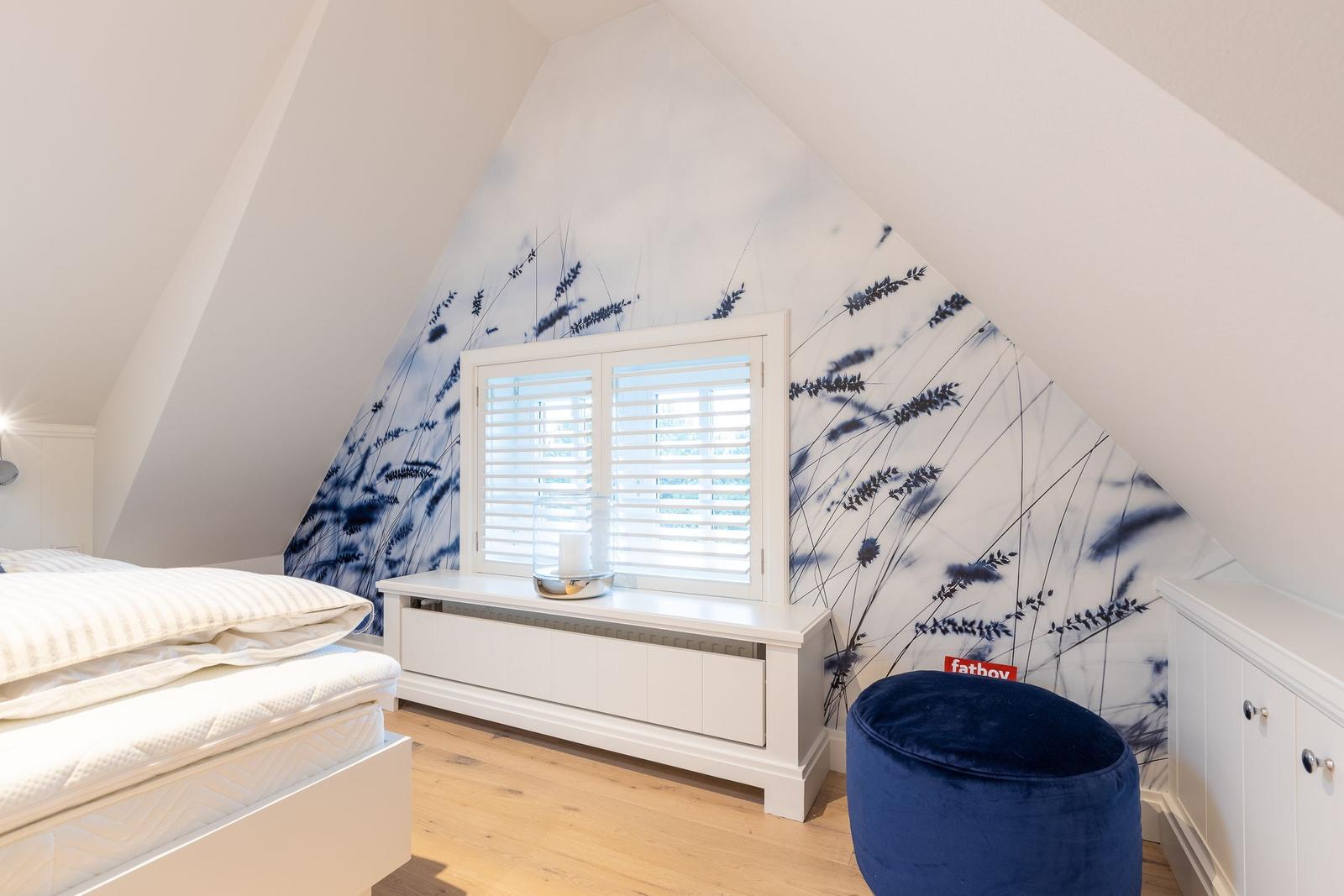 Schlafzimmer mit Dachschrägen, Fenster mit Jalousie und blauem Sitzkissen.