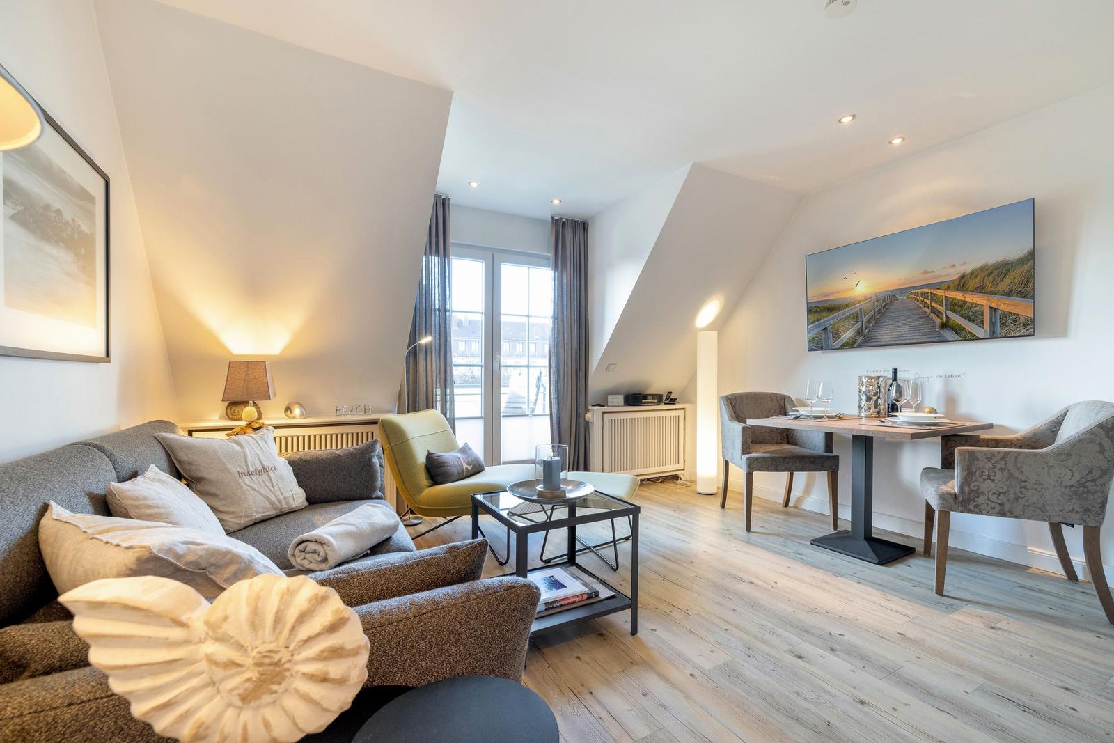 Living and dining area with sofa, chair, and table. Wooden floor and large windows.