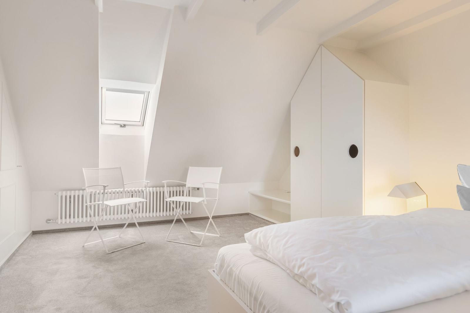 Schlafzimmer mit Dachschrägen, weißem Bett, Stühlen und Fenster.