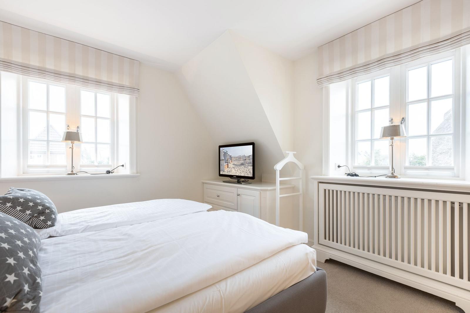 Schlafzimmer mit Doppelbett, zwei Fenstern und Fernseher auf Schrank.