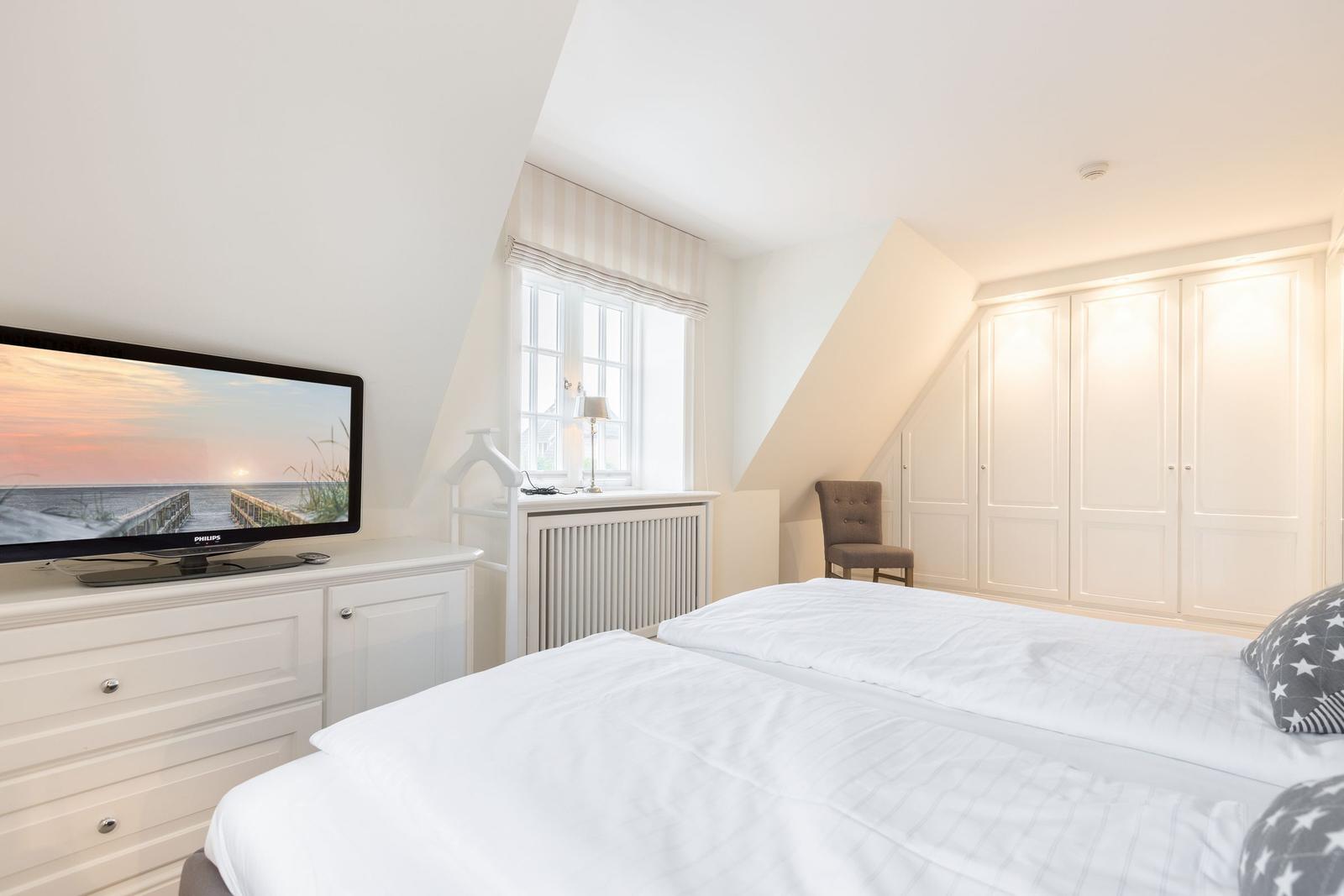 Schlafzimmer mit Bett, TV und Kleiderschrank. Fenster mit Vorhang und Stuhl.