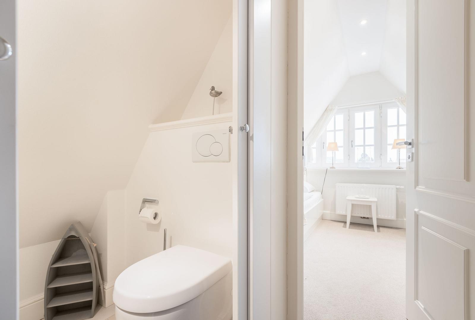 Badezimmer mit Toilette und kleinem Regal. Blick in Schlafzimmer mit Fenster und Stuhl.