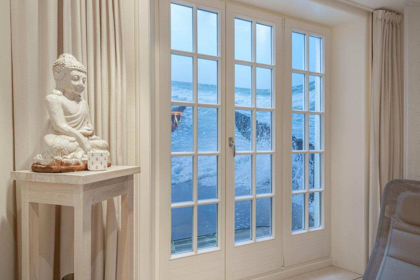Durch die Fenster sieht man das Meer mit Wellen. Eine Buddha-Statue steht auf einem Tisch.