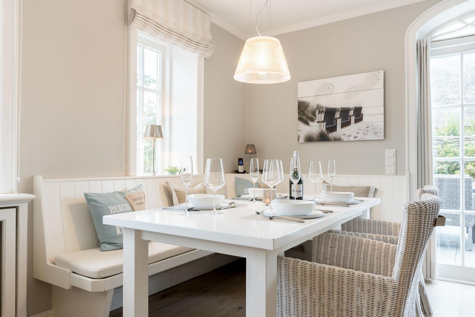 Esszimmer mit weißem Tisch, Bänken und Stühlen, Fenstern und Wandbild.