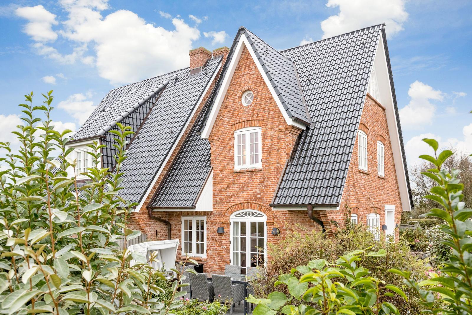 Rotes Backsteingebäude mit dunklem Satteldach und weißen Fenstern. Gartenbereich mit Sitzgelegenheit.