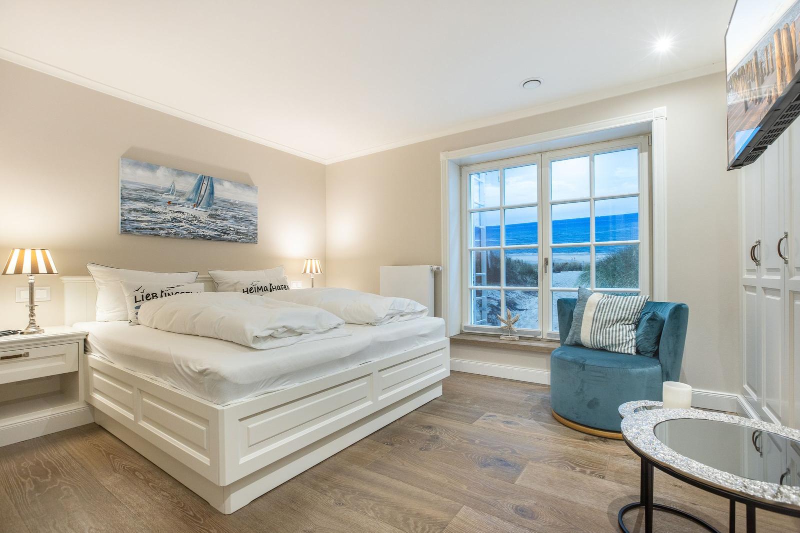 Schlafzimmer mit Blick aufs Meer, weißes Bett, blauer Sessel und Fenster mit Meerblick.