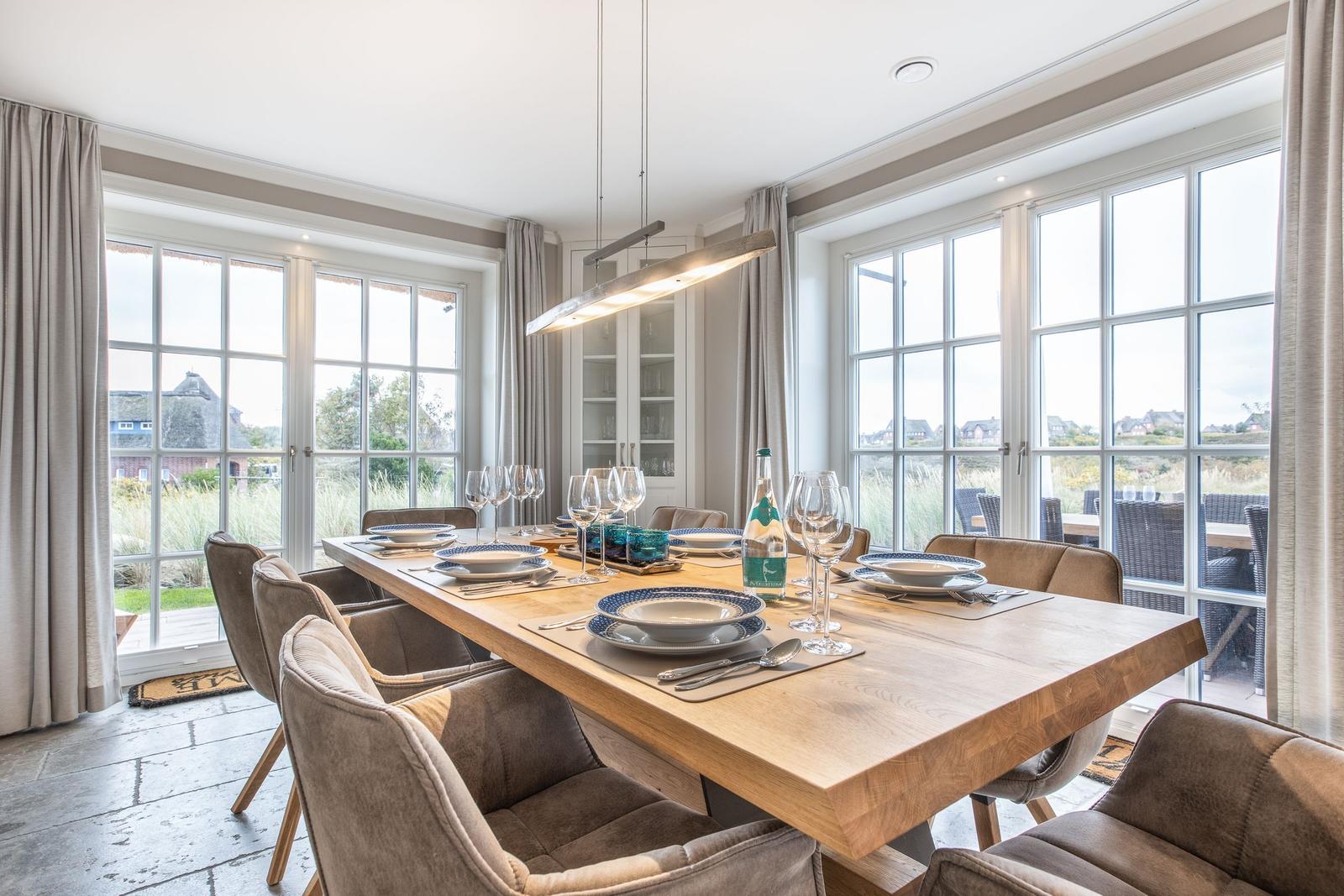 Esszimmer mit langem Holztisch, Stühlen und großen Fenstern mit Blick nach draußen.