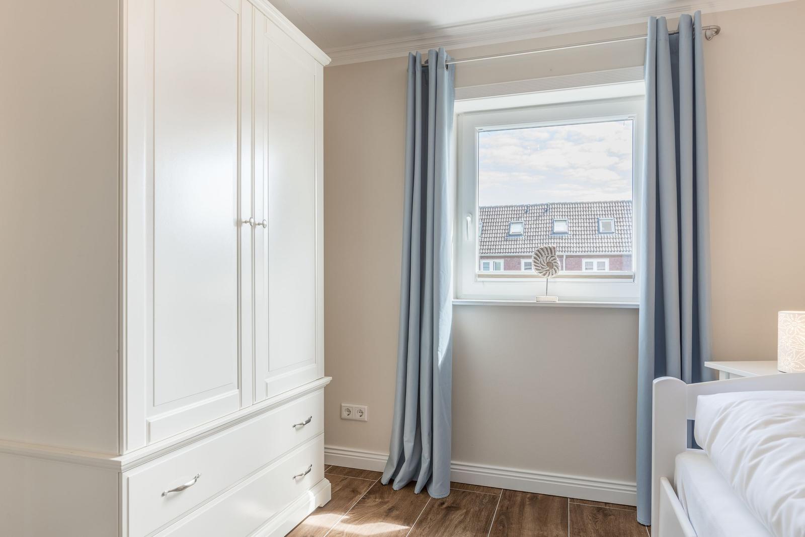 Schlafzimmer mit weißem Schrank, Fenster mit Vorhängen und Blick auf Dach