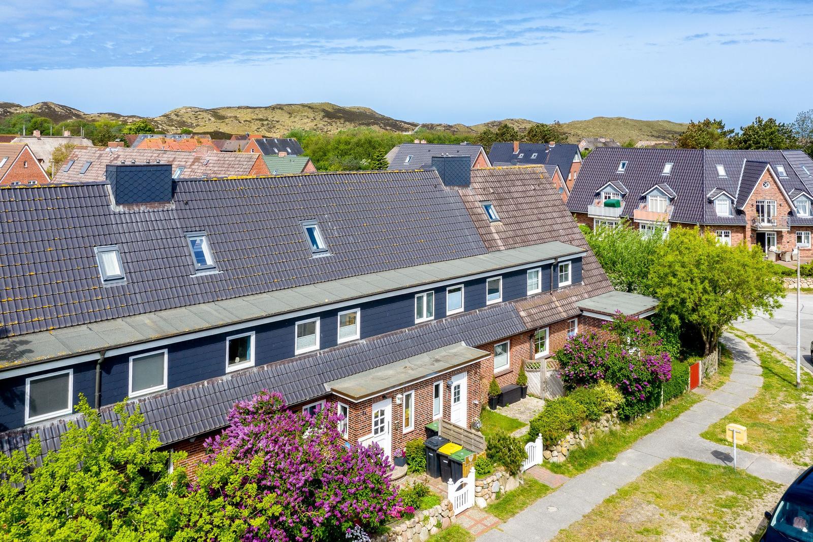 Haus mit dunkelblauem Dach und roten Ziegelsteinen, umgeben von grünen Bäumen und lila Blumen.