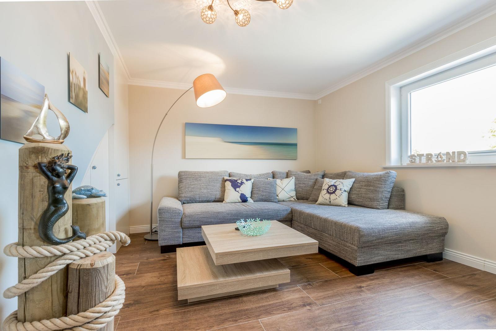 Wohnzimmer mit Sofa, Couchtisch und Seemotivskulptur. Fenster mit Strand-Schriftzug.