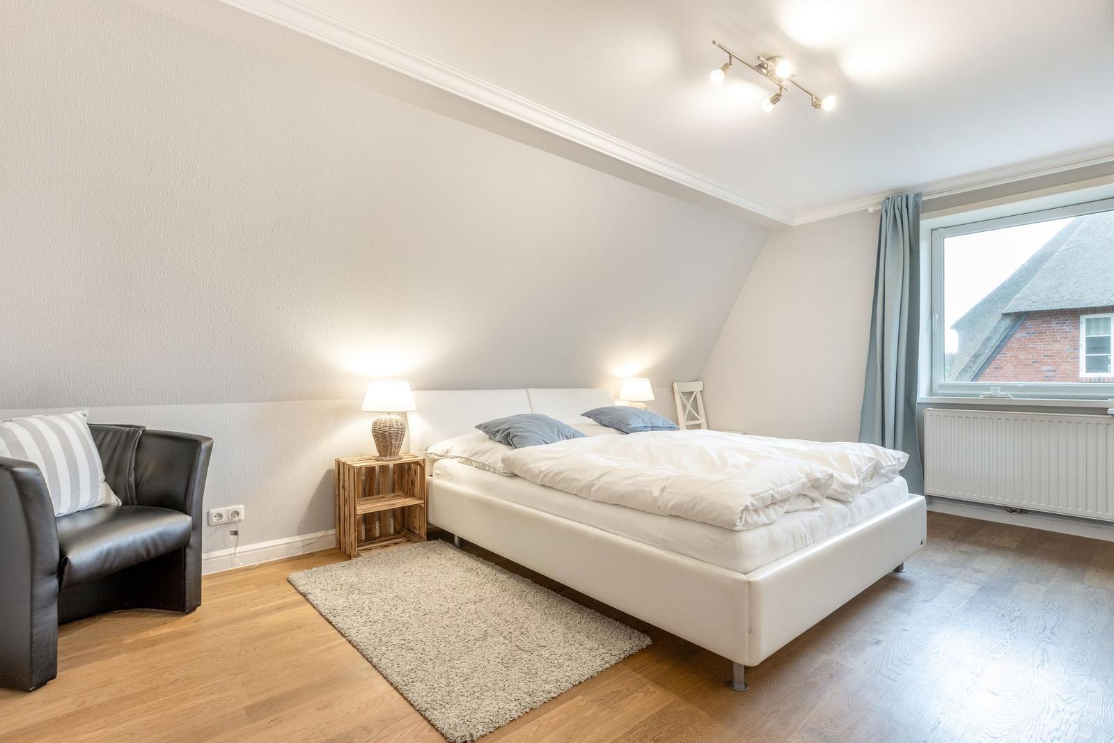 Bedroom with bed, armchair, and window. Wooden floor and white walls.