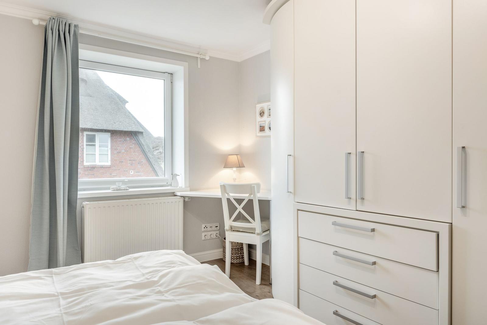 Bedroom with white desk, chair, and wardrobe. Window shows thatched roof house.