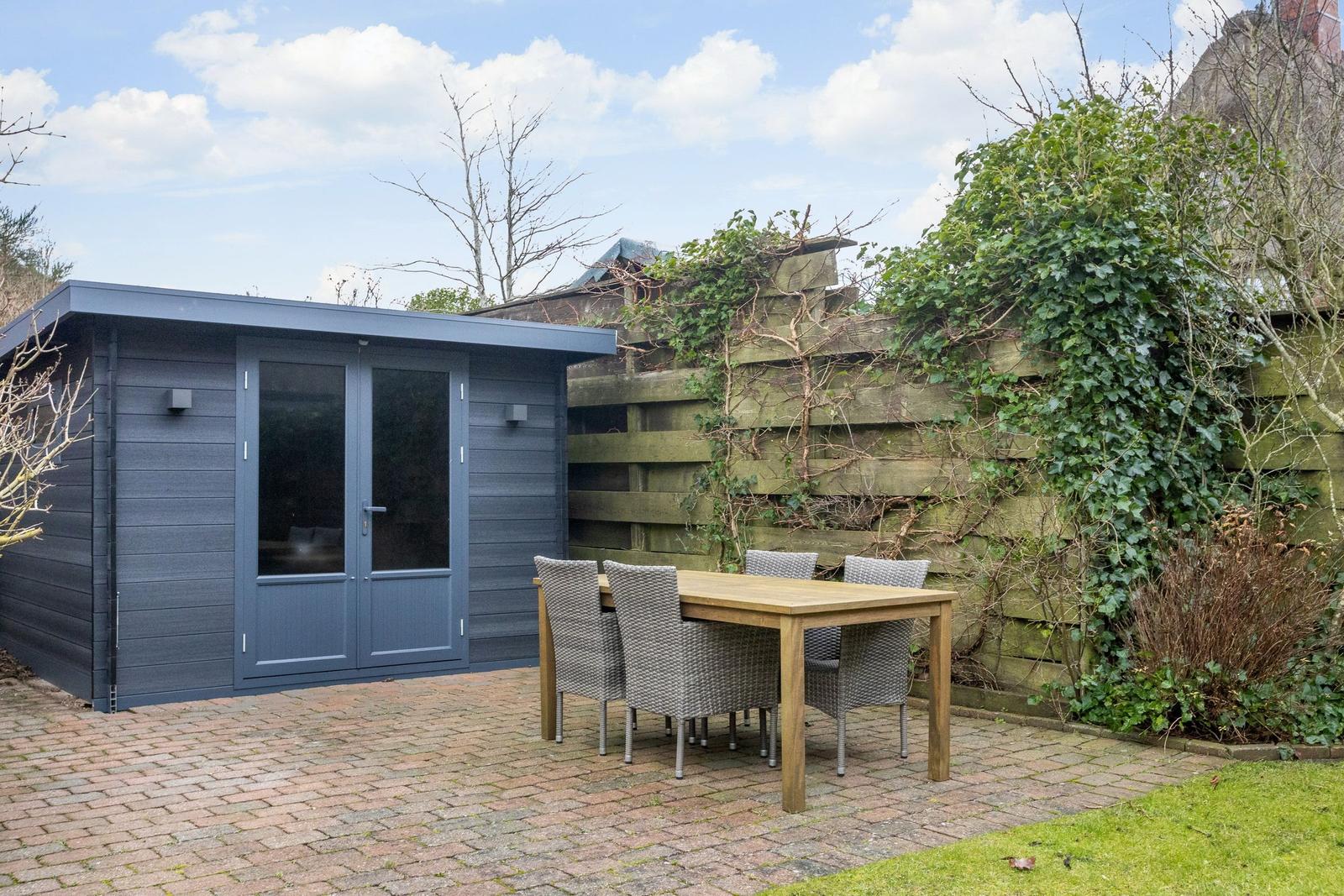 Garden house with terrace, table and chairs. Wooden fence with ivy. Paved path and grass area.