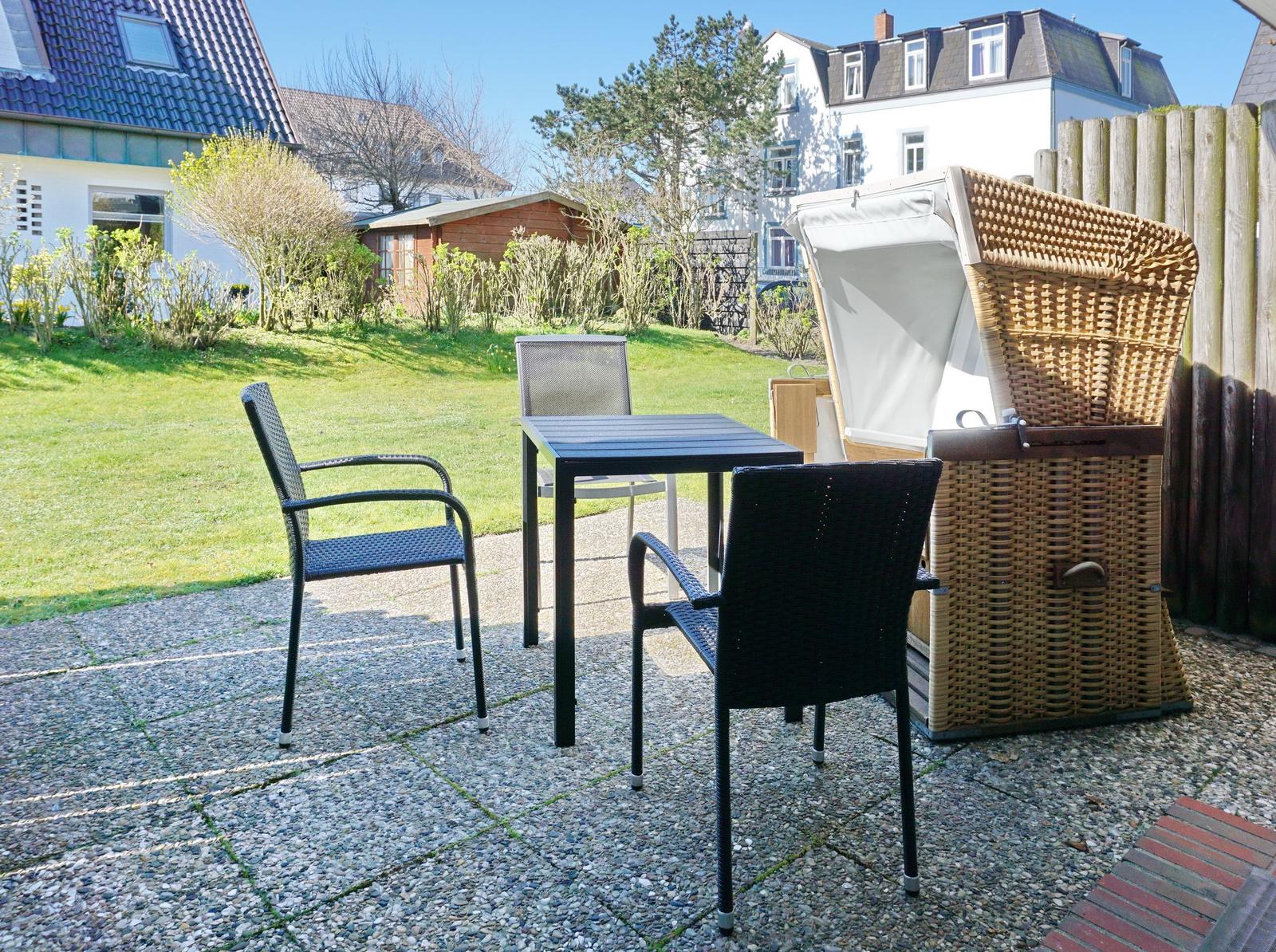 Terrace with table, three chairs, and lounge chair. Background: garden and houses.