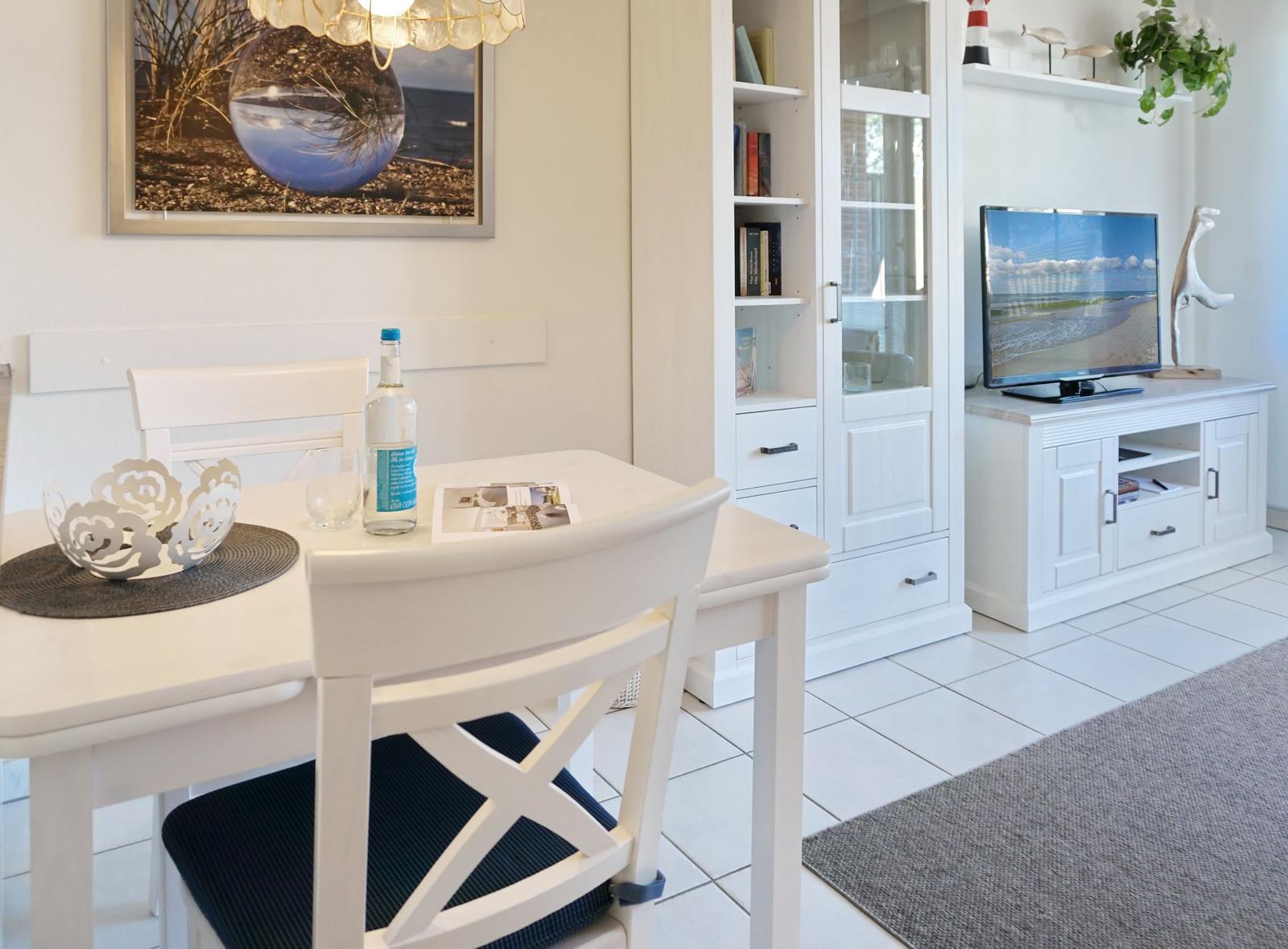 Dining area with white table and chairs, TV and bookcase in background.