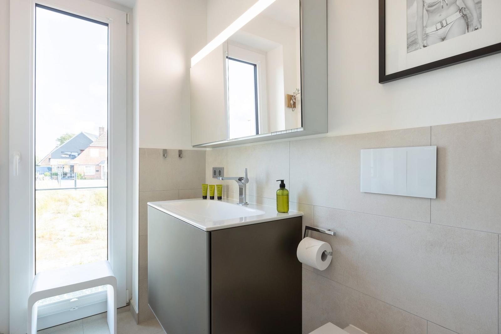 Badezimmer mit Waschbecken, Spiegel und Fenster. Wandverkleidung und Toilettenpapierhalter sichtbar.