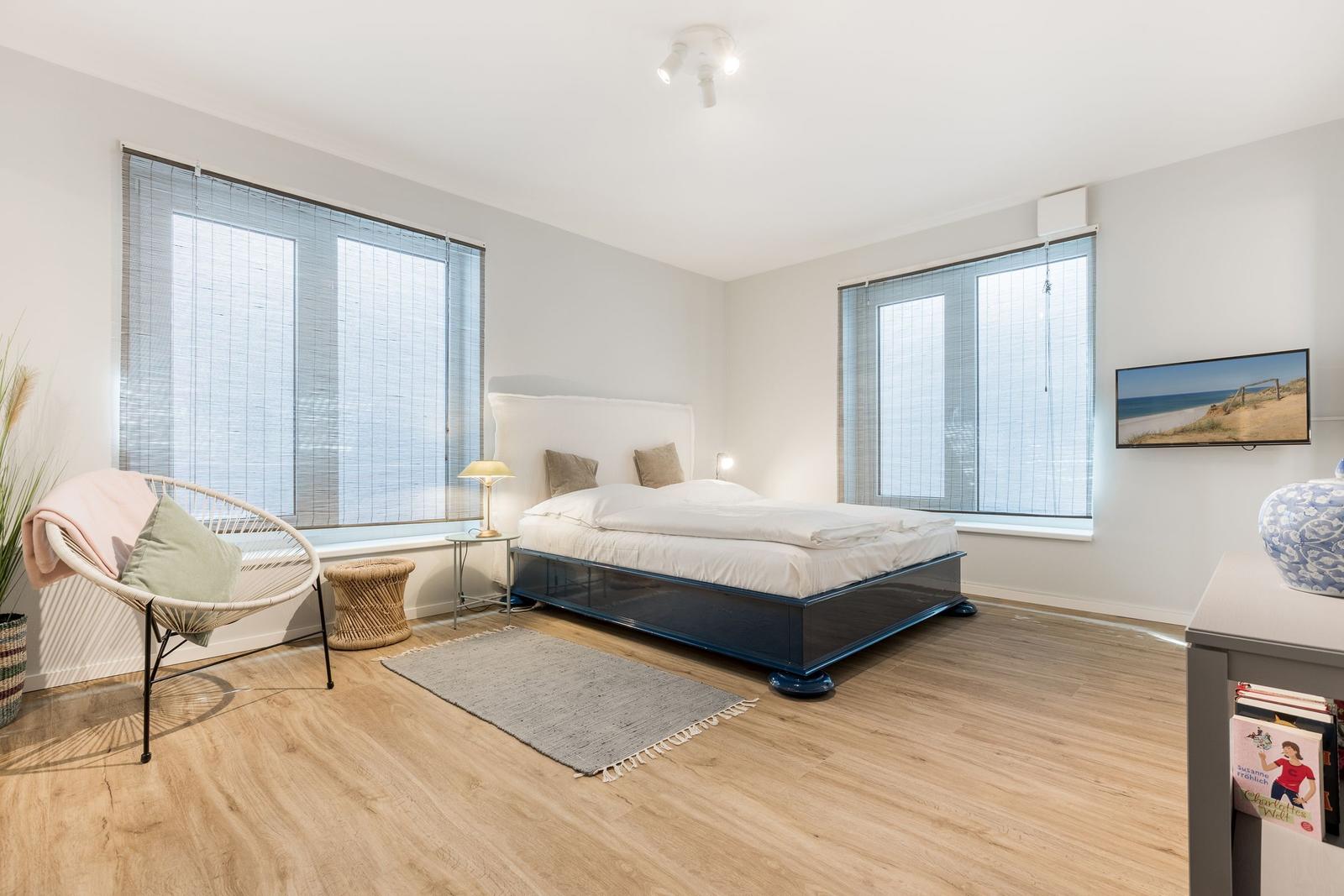 Schlafzimmer mit Bett, Fenstern und Sitzgelegenheit.